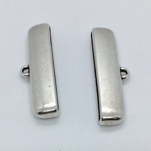 Op de afbeelding: Twee zilverkleurige metalen schuifgespen met een lus aan elk uiteinde. De gespen zijn rechthoekig en hebben een gladde, gepolijste afwerking.