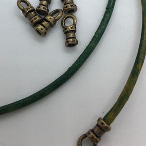 May include: Five gold-toned metal cord ends with a loop on one end and a decorative design on the other. The cord ends are arranged on a white surface next to a green leather cord.