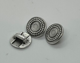 May include: Three silver buttons with a decorative design. The buttons have a raised, circular pattern with a smaller circle in the center. One button has a loop on the back.