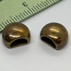 May include: Two antique brass-colored metal half-dome beads with a curved, open base. The beads have a slightly textured surface and a warm, aged appearance. A green ruler is partially visible in the background, providing scale.