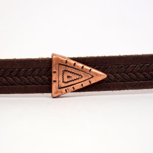 Puede incluir: Una correa de cuero marrón con una hebilla triangular de cobre. La hebilla tiene un diseño geométrico.