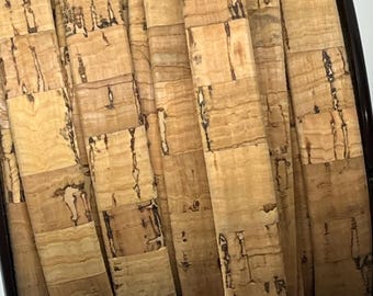 May include: Close-up of a roll of cork material. The cork has a natural, light brown color with darker brown and black markings. The cork is arranged in a rectangular pattern, creating a textured surface. The roll is contained within a dark brown border.