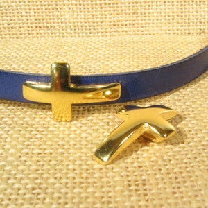 Op de afbeelding: Twee gouden kruisbedels, een bevestigd aan een blauw leren armband. De bedels zijn eenvoudig en elegant, met een gladde, gepolijste afwerking.