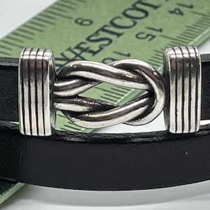 May include: A black leather bracelet with a silver knot-shaped clasp. The clasp has a braided design.
