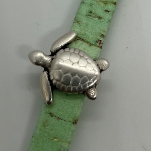May include: A silver turtle charm with a detailed shell design, attached to a green cork bracelet.
