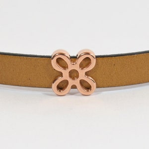 May include: A rose gold metal charm in the shape of a flower with four petals. The charm is attached to a brown leather strap.