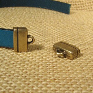 May include: Two gold-toned metal clasps for a leather strap. One clasp is closed and the other is open. The clasps are on a brown burlap background.