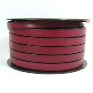 May include: A spool of burgundy leather strips, tightly wound. The leather appears smooth with a dark edge, creating a contrast. The spool is set against a white background, highlighting the rich colour and texture of the material.