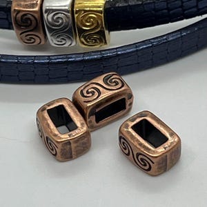 May include: A bracelet featuring decorative metal beads in copper, silver, and gold, with an engraved swirl pattern. The bracelet has a dark blue leather strap. The beads are rectangular.