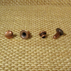May include: Four copper rivets of different sizes and styles. The rivets are on a brown burlap background.