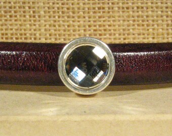 May include: A silver-toned round charm with a clear crystal in the center. The charm is on a brown leather background.
