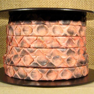 May include: A stack of six decorative bracelets with a snakeskin pattern in shades of pink and gray. The bracelets are stacked on a black circular base, with a black top. The background is a tan burlap.