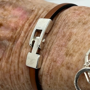 May include: A brown leather bracelet with a silver clasp. The clasp is a simple design with a rectangular shape and a small round accent.