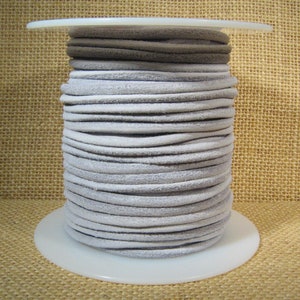 May include: A spool of light gray suede cord, wound tightly on a white plastic spool. The cord is approximately 3 mm in diameter.