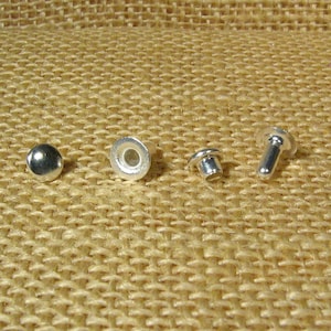 May include: Four silver metal rivets, two with a flat head, one with a round head, and one with a post. The rivets are on a brown burlap background.
