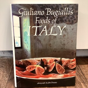 May include: A hardcover cookbook titled "Giuliano Bugialli's Foods of ITALY" with a photograph of figs and prosciutto. The cover features a photo of figs and prosciutto on a plate. The book is on a wooden surface.