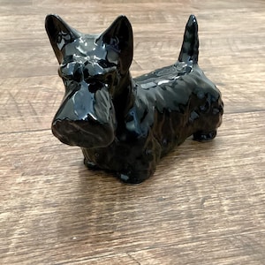 May include: A glossy black ceramic figurine of a Scottish Terrier dog. The dog has upright ears, a long snout, and a short tail. The figurine is sitting on a wooden surface.
