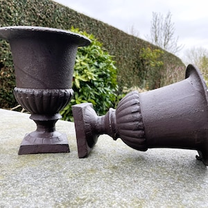 Puede incluir: Dos macetas de jardín de hierro fundido de color marrón oscuro, una en posición vertical y la otra tumbada de lado. Las macetas tienen una forma de urna clásica con un diseño acanalado alrededor de la parte superior.