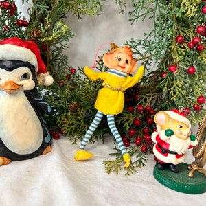 May include: Three Christmas ornaments: a penguin wearing a Santa hat, a yellow elf with blue and white striped legs, and a mouse wearing a Santa suit holding a mirror.