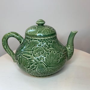 Może przedstawiać: Szałwiowo-zielony ceramiczny czajniczek z wypukłym wzorem liści winorośli, pnączy i winogron pokrywającym cały korpus oraz pokrywkę. Czajniczek posiada zakrzywiony uchwyt, zwężający się dzióbek oraz dopasowaną pokrywkę z małym, okrągłym uchwytem.