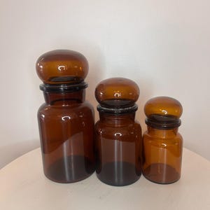 Könnte beinhalten: Drei bernsteinfarbene Apothekergläser in unterschiedlichen Höhen, jedes mit einem zylindrischen Körper und einem passenden, abgerundeten, kuppelförmigen Glasdeckel, der sicher auf dem Rand des Glases sitzt und ein einheitliches Set bildet.