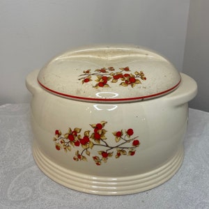 May include: A white ceramic covered dish with a red and gold floral design. The dish has a round shape and a lid with a small handle on each side.