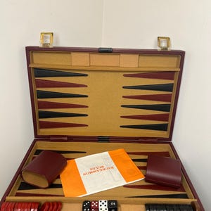 Puede incluir: Un juego de backgammon de cuero burdeos con un tablero de madera, dados, fichas y un libro de reglas. El juego está abierto y listo para jugar.