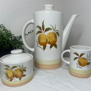 Puede incluir: Un juego de té de cerámica blanca con un diseño de fruta amarilla y verde. El juego incluye una tetera, una jarra de crema y un azucarero.