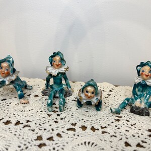 May include: Four ceramic figurines of whimsical elves with green and white painted details. The elves are in various poses, some sitting, some crawling, and some standing. They are all wearing green and white hats and have painted facial features.