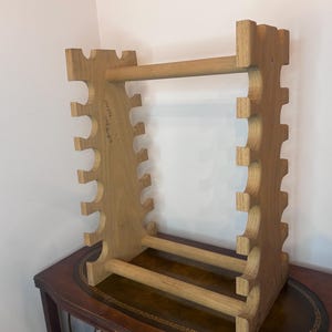 May include: A light brown wooden wine glass rack with a rectangular frame. The rack has a series of curved cutouts along the sides to hold glasses, and horizontal bars for support. The rack is sitting on a dark brown table.