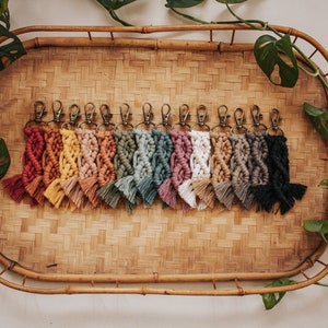 May include: A collection of macrame keychains in various colors, including red, yellow, pink, green, white, and black. Each keychain features a woven design and a metal clasp. The keychains are displayed on a woven tray.
