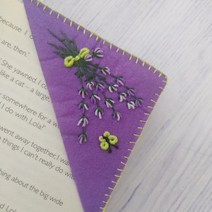 Puede incluir: Un marcapáginas triangular de fieltro morado con un bordado de un ramo de lavanda y una pequeña flor. Los bordes están cosidos con hilo amarillo. El marcapáginas está colocado en un libro.