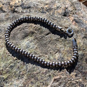 K&ouml;nnte beinhalten: Ein silbernes Armband mit Perlen und einem Karabinerverschluss. Das Armband liegt auf einer rauen, grauen Steinoberfl&auml;che.