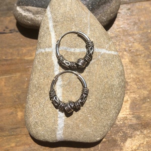 Peut inclure: Une paire de boucles d'oreilles créoles en argent avec des détails complexes. Les boucles d'oreilles présentent des éléments décoratifs, notamment du fil torsadé et des formes stylisées. Elles sont posées sur une pierre claire.