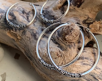 May include: Silver hoop earrings with decorative beaded detailing. The earrings are presented on driftwood, highlighting their design. The hoops are different sizes, offering a versatile jewellery option. The earrings are made of silver.