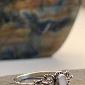 May include: A silver ring with an oval, white gemstone set in an ornate, filigree design. The ring is on a gray surface, with a blurred background of a ceramic vase.