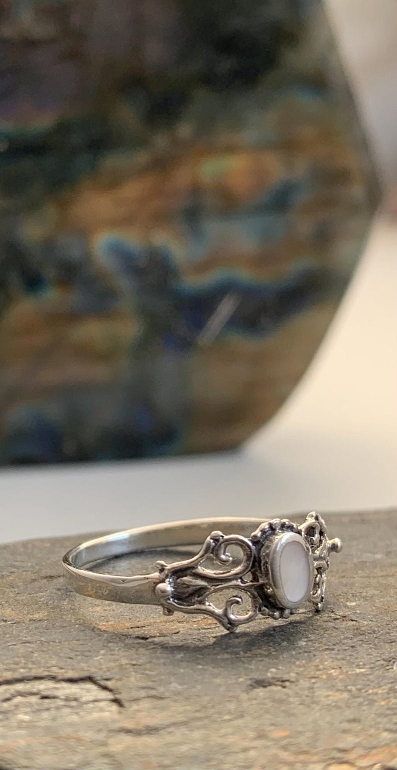 May include: A silver ring with an oval, white gemstone set in an ornate, filigree design. The ring is on a gray surface, with a blurred background of a ceramic vase.