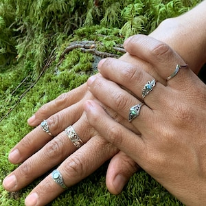May include: Close-up of hands adorned with several silver rings. The rings feature intricate designs, including filigree and gemstone settings. The background is a bed of vibrant green moss, creating a natural and earthy aesthetic.