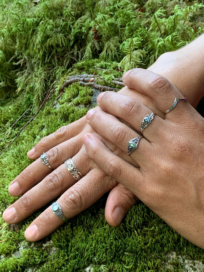 May include: Close-up of hands adorned with several silver rings. The rings feature intricate designs, including filigree and gemstone settings. The background is a bed of vibrant green moss, creating a natural and earthy aesthetic.