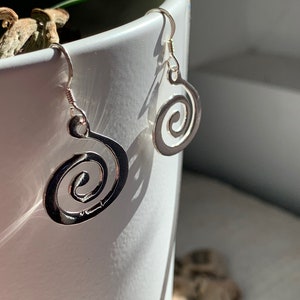 Op de afbeelding: Een paar zilveren oorbellen met een spiraalvormig ontwerp. De oorbellen hangen aan een witte keramische pot.