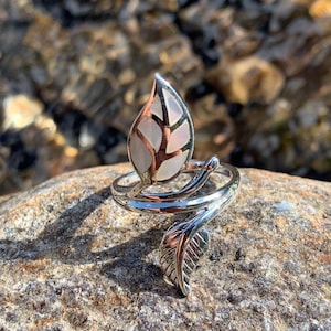 Könnte beinhalten: Ein silberfarbener Ring mit Blattdesign. Der Ring hat ein großes blattförmiges Mittelteil mit einem weißen und rosa Muster. Ein kleineres Blattdetail ziert das Band, das sich um den Finger wickelt. Der Ring ist vor einem Naturstein-Hintergrund platziert.