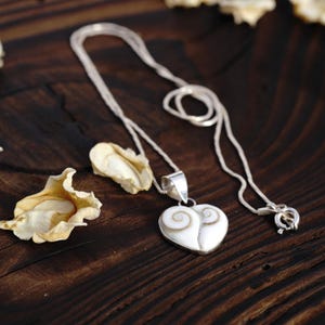 Op de afbeelding: Een zilveren ketting met een hartvormige hanger gemaakt van twee stukken wit en bruin spiraalschelp. De hanger is bevestigd aan een gepolijst zilveren oogje en de ketting is voorzien van een standaard zilveren veerringsluiting.