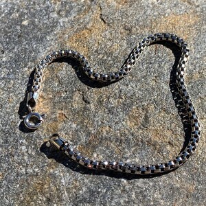 May include: A silver chain bracelet with a box chain design. The bracelet is lying on a grey textured surface.