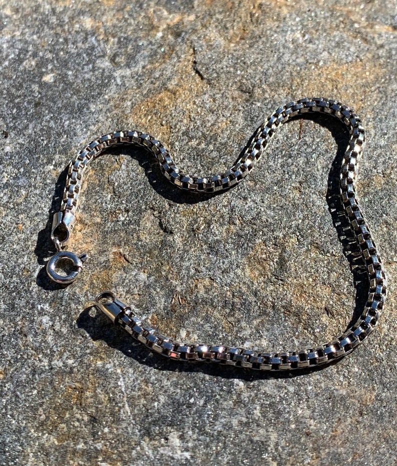 May include: A silver chain bracelet with a box chain design. The bracelet is lying on a grey textured surface.