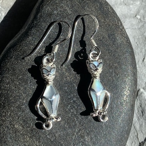 May include: A pair of silver cat earrings with iridescent mother-of-pearl inlay. The earrings feature a stylized cat design with detailed paws and a curved tail. The hooks are made of silver and are designed for pierced ears.
