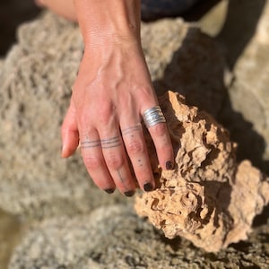 K&ouml;nnte beinhalten: Nahaufnahme einer Hand, die am Ringfinger einen Stapel silberner Ringe tr&auml;gt. Die Hand ruht auf einem St&uuml;ck rauen, braunen Gesteins.