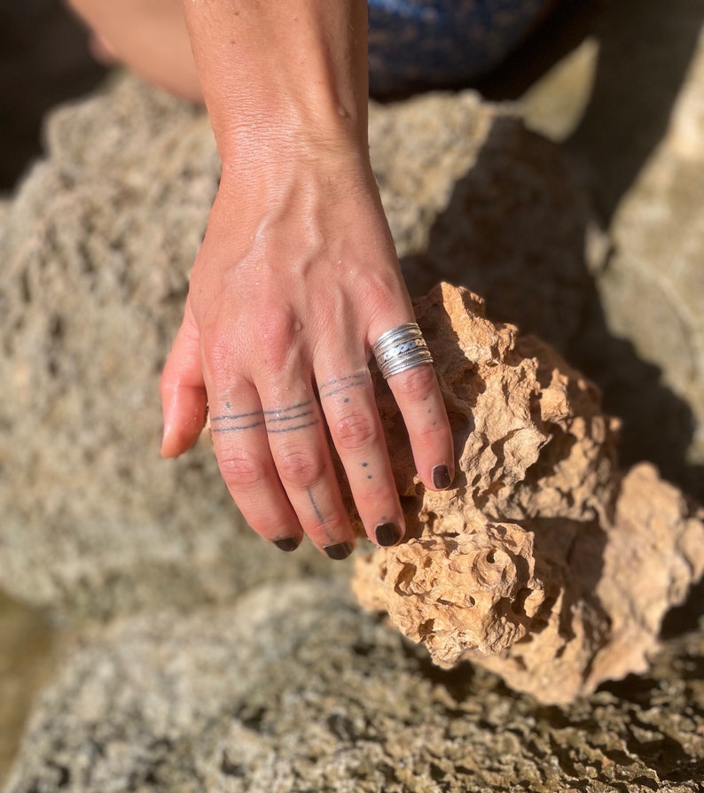 K&ouml;nnte beinhalten: Nahaufnahme einer Hand, die am Ringfinger einen Stapel silberner Ringe tr&auml;gt. Die Hand ruht auf einem St&uuml;ck rauen, braunen Gesteins.