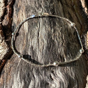 Puede incluir: Una delicada pulsera de plata con una serie de pequeñas cuentas redondas intercaladas a lo largo de una fina y flexible cadena. La pulsera se asegura con un pequeño cierre. La joya se exhibe sobre una superficie de madera oscura texturizada.