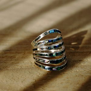 May include: A multi-band ring featuring five silver-toned curved bands. Each band is inlaid with small, rectangular pieces of iridescent abalone shell that display shifting hues of blue, green, and purple against the metallic silver finish.