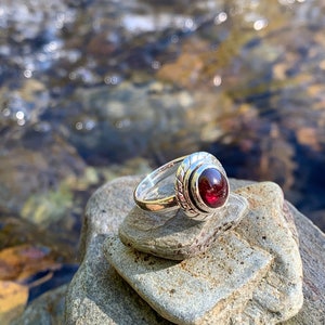Peut inclure: Une bague en argent avec une pierre précieuse rouge foncé sertie dans une bélière en argent décorative. La bague est posée sur une pile de pierres grises.